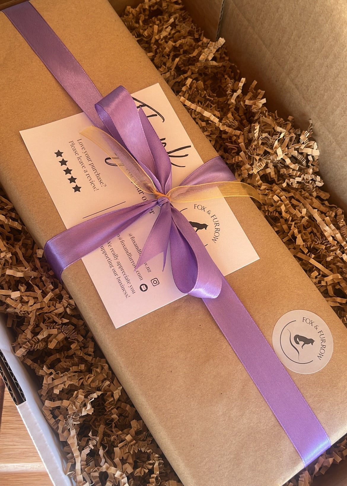 A brown kraft paper gift box wrapped with a purple ribbon and a white card, placed inside a larger cardboard box filled with shredded paper filler.