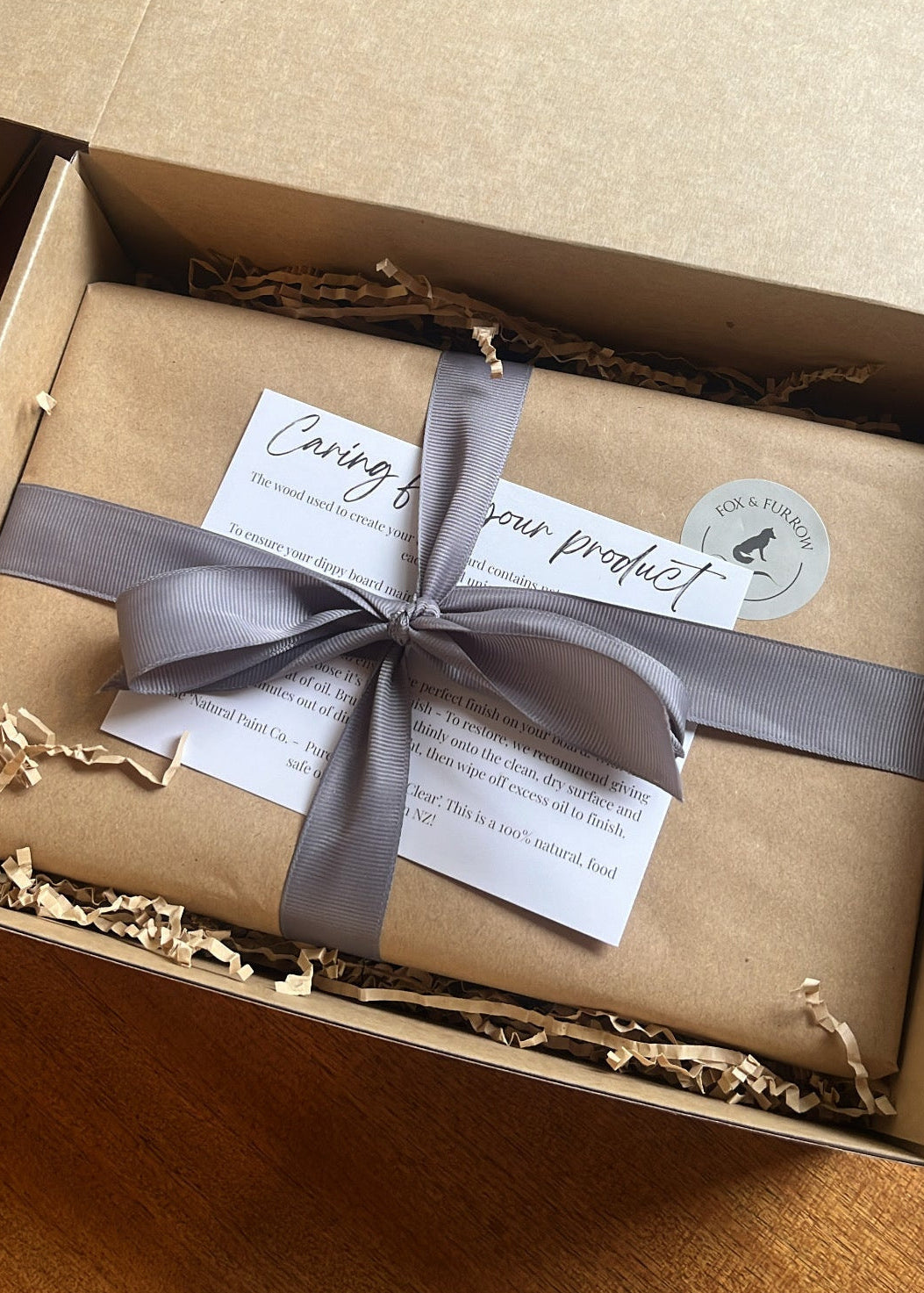 A gift box containing a brown kraft paper wrapped package tied with a gray ribbon and a white card, shown inside an open brown cardboard box on a wooden surface.