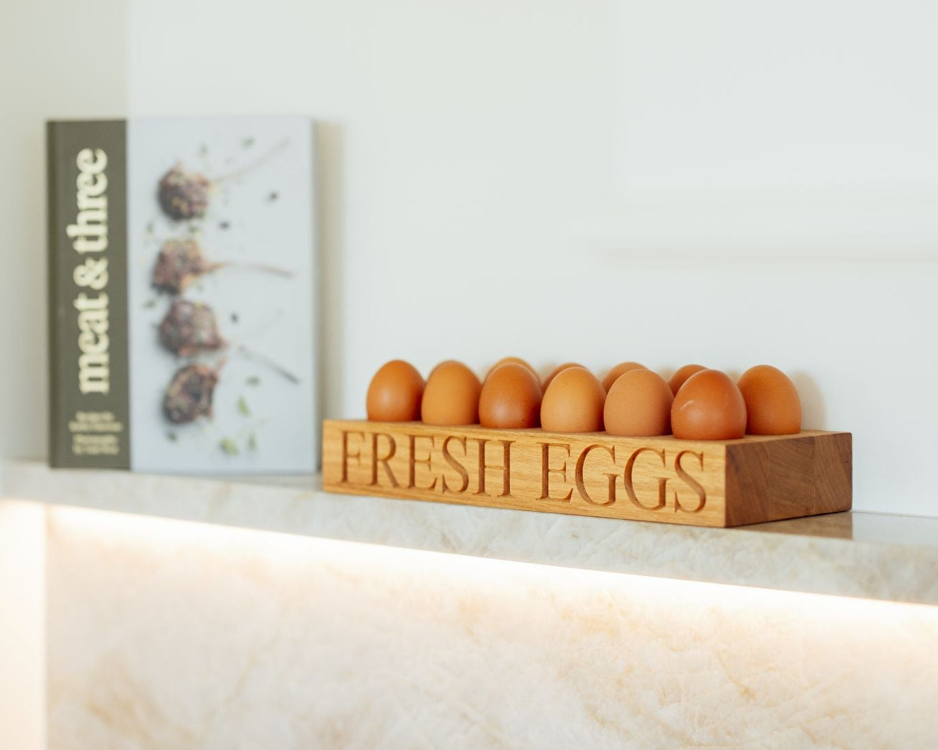 Wooden egg holder with eggs on a kitchen counter next to a cookbook titled 'meat & three'.