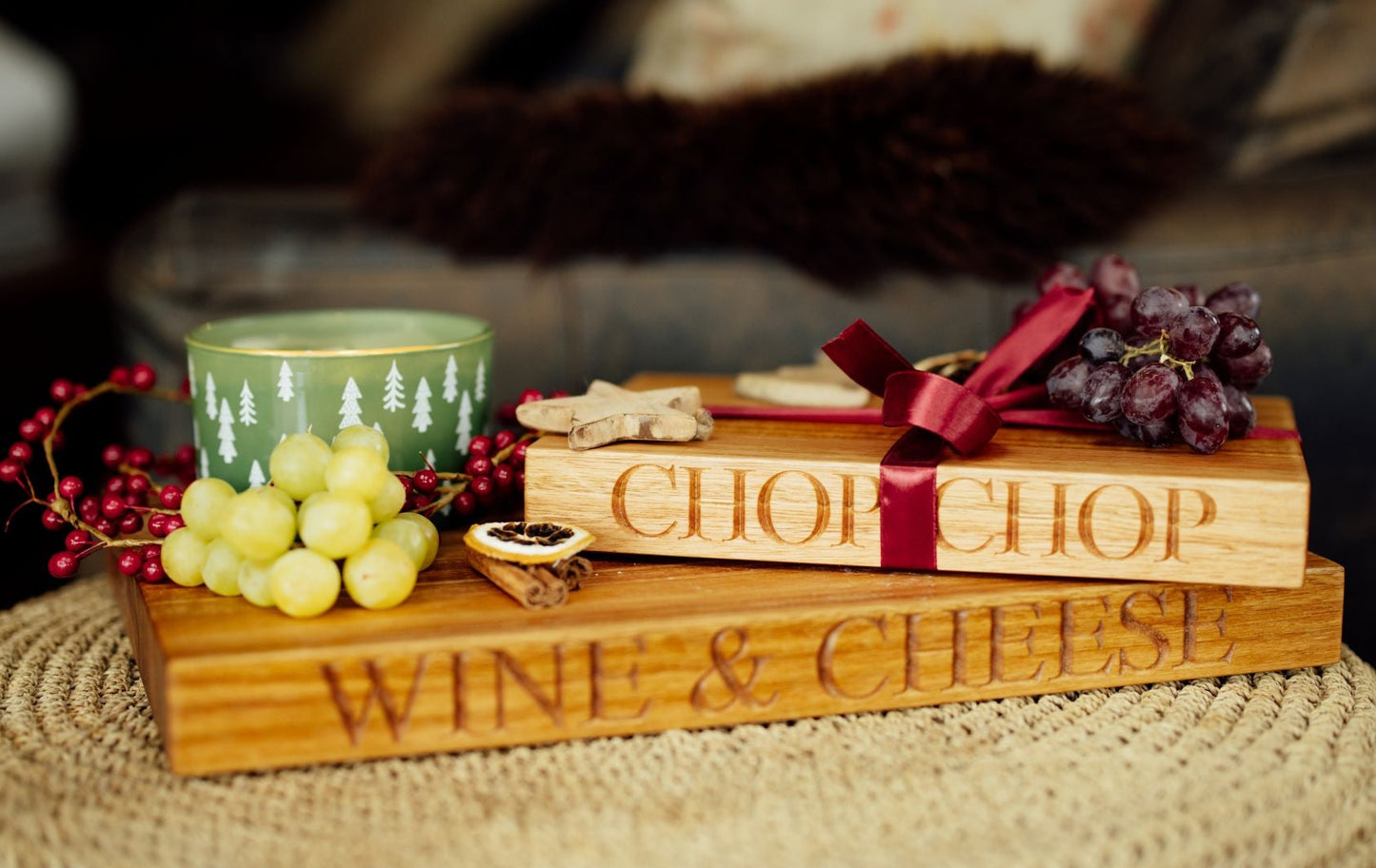 A styled gift set on a woven ottoman featuring two engraved wooden blocks reading 'CHOP CHOP' and 'WINE & CHEESE,' decorated with grapes, a red ribbon, and a green candle, in a softly blurred indoor setting.