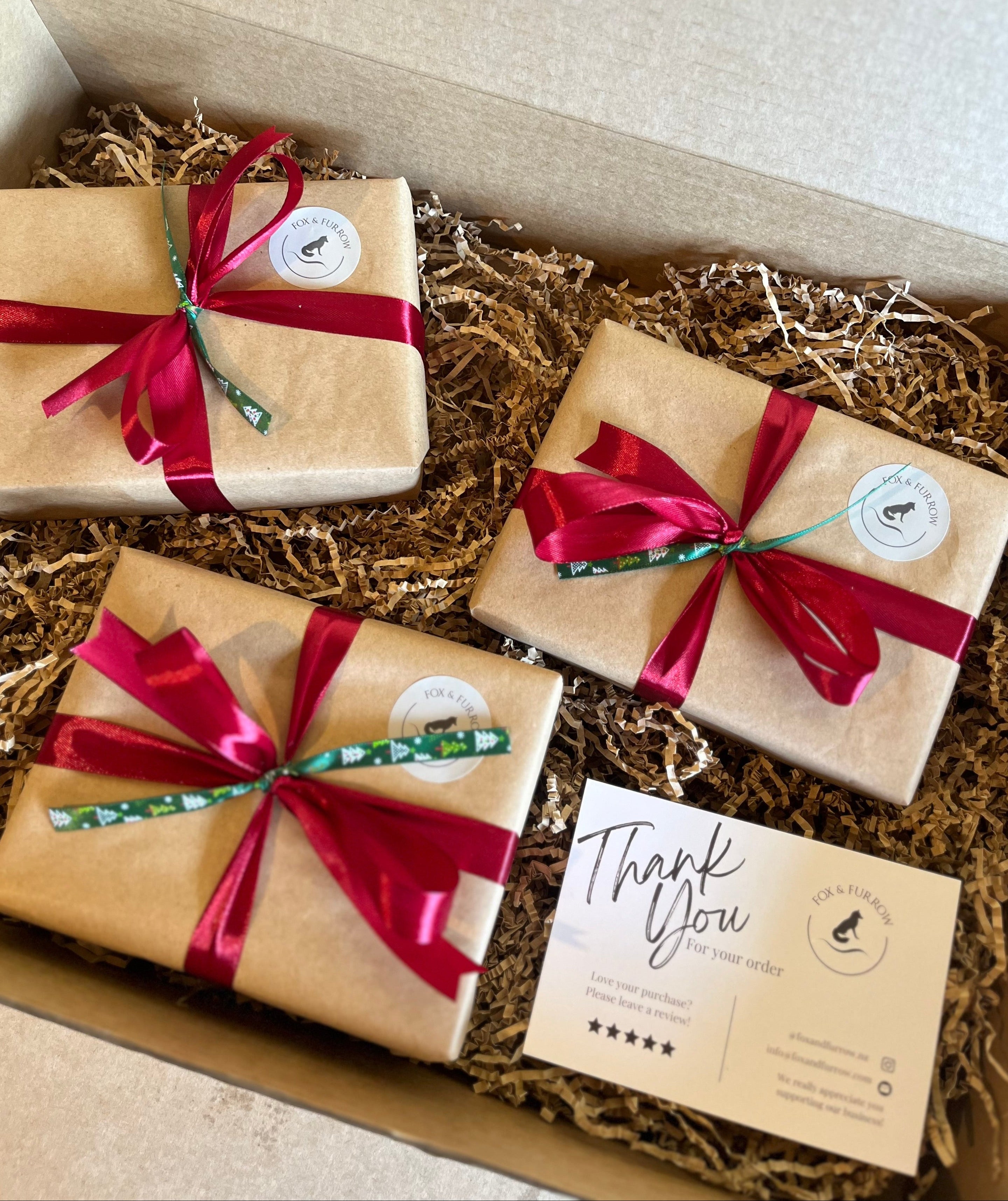 Three wrapped gift boxes in a cardboard box filled with shredded paper, each tied with a red ribbon and a small green patterned accent strip. A beige thank-you card is placed in the box.