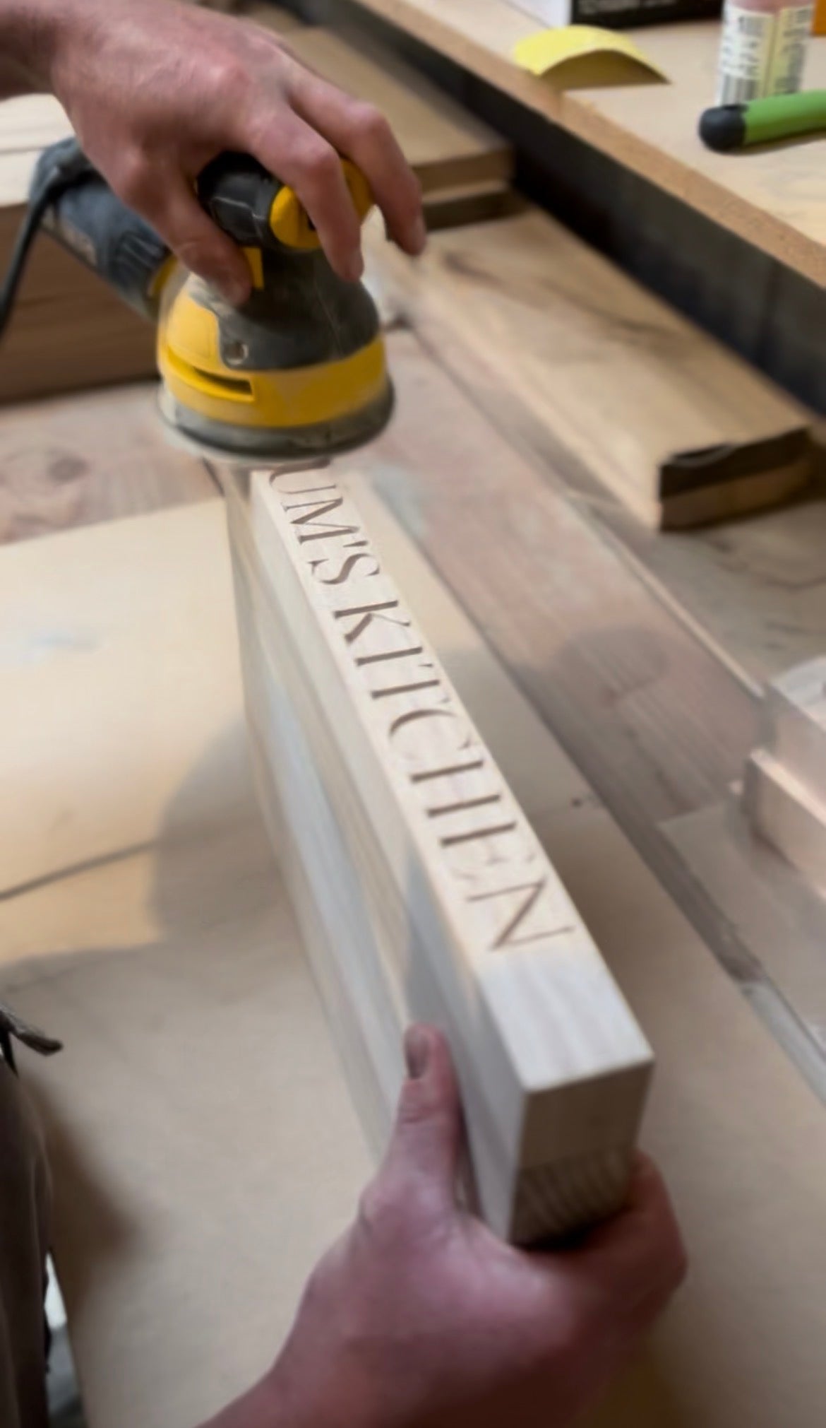 Person sanding a wooden chopping board with 'Mum's kitchen' engraved into it, on a workshop table.