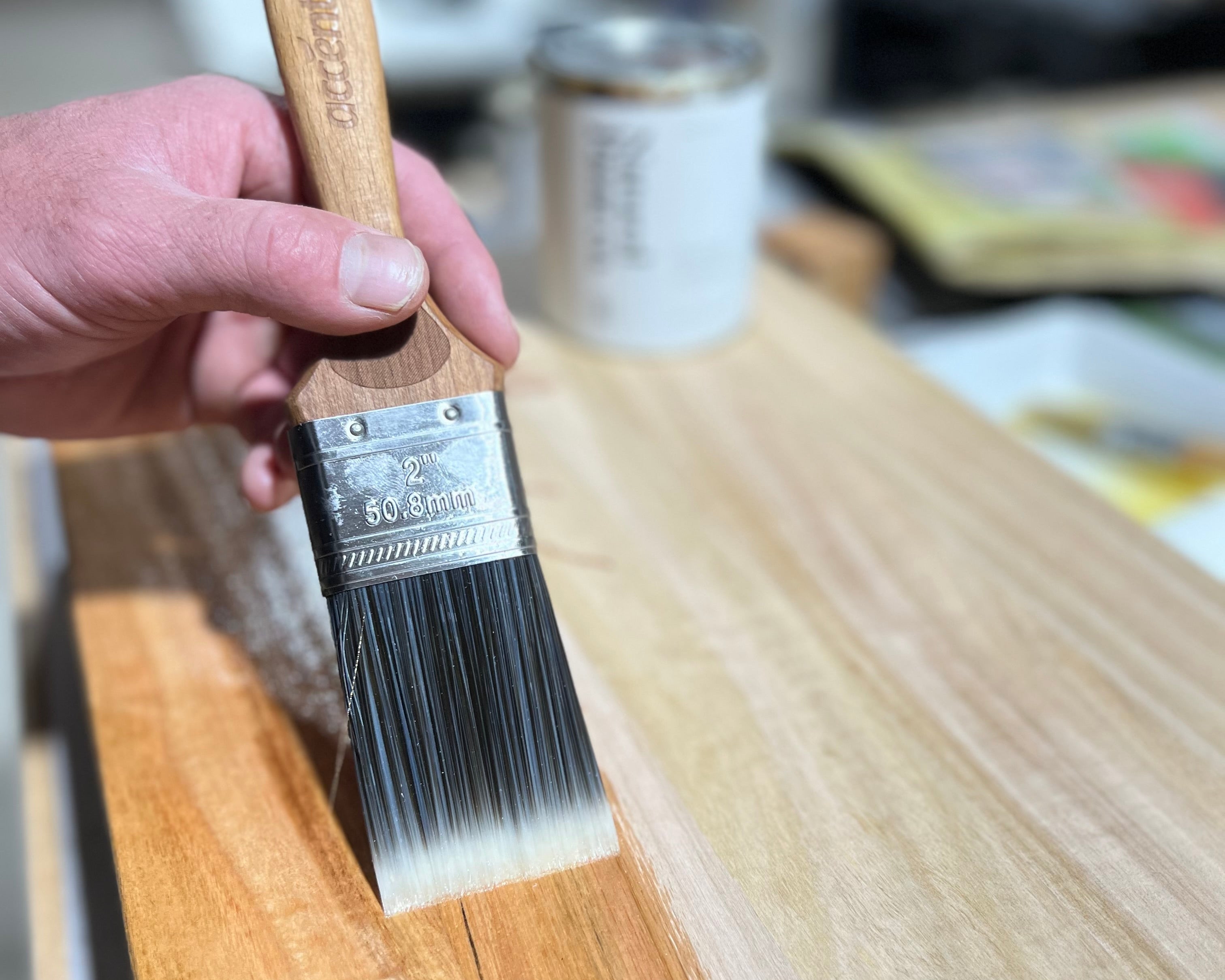 Person holding a paintbrush with a wooden handle and metal ferrule, applying paint to a wooden surface.