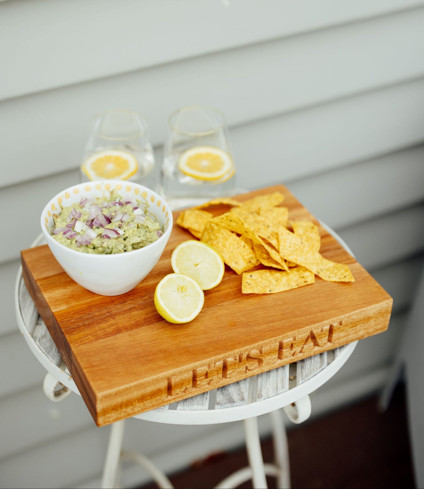 A wooden serving board engraved with 'LET'S EAT' sits on a white table, styled with tortilla chips, a bowl of guacamole, lemon slices, and two glasses of iced coffee against a light gray siding background.