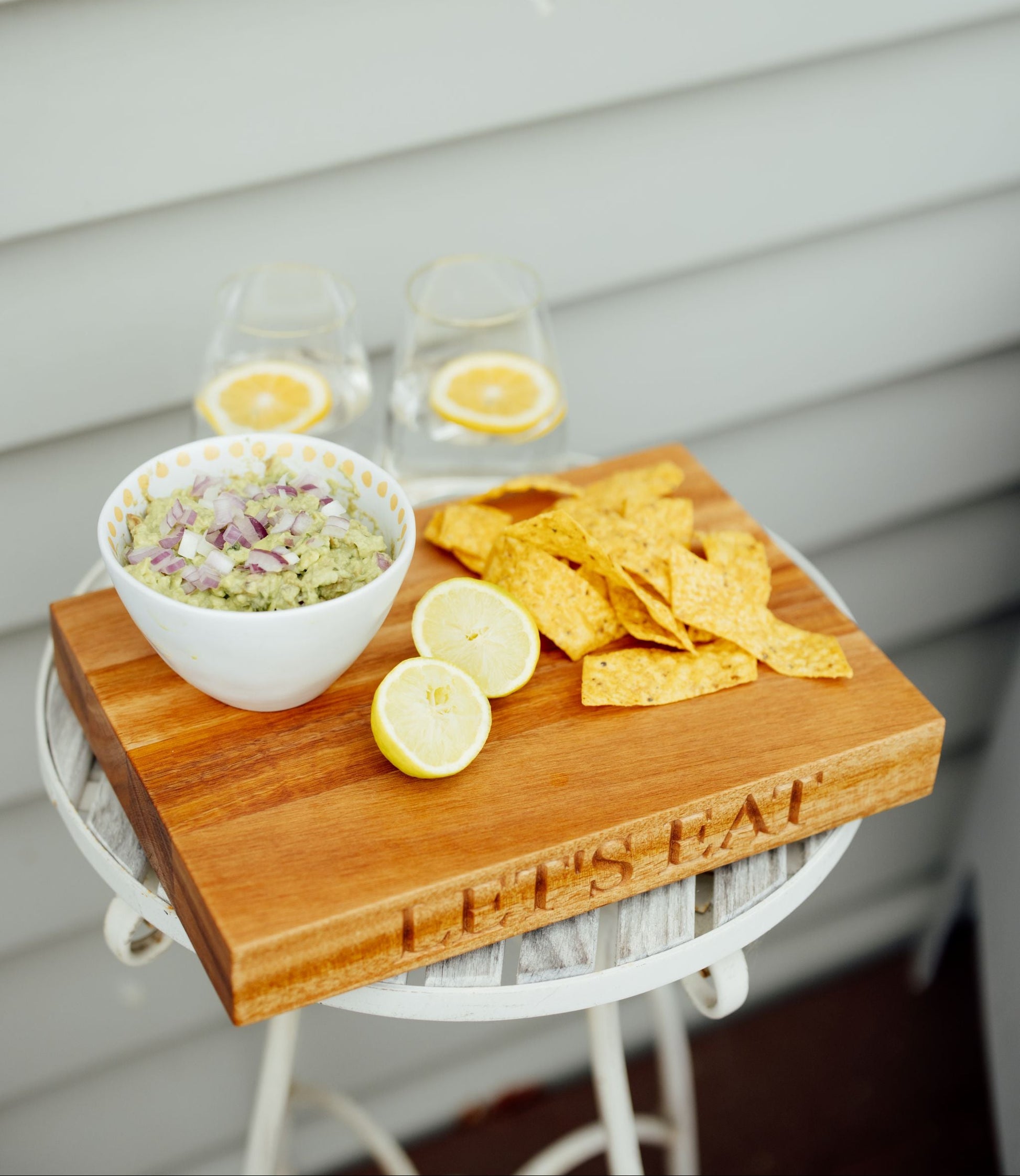 A wooden serving board engraved with 'LET'S EAT' sits on a white table, styled with tortilla chips, a bowl of guacamole, lemon slices, and two glasses of iced coffee against a light gray siding background.
